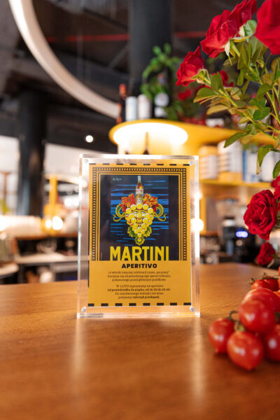 Plakat reklamowy Martini Aperitivo ustawiony na drewnianym barze restauracji LUPO – kampania promująca włoską kulturę aperitivo, z winogronami i butelką trunku na grafice vintage. Martini x Mateo Zielonka