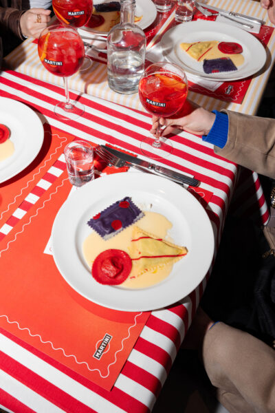 Mateo Zielonka – szef kuchni znany jako Pasta Man – uśmiecha się, prezentując kolorowy talerz ravioli, stojąc pod szyldem „Martini Aperitivo” w nowoczesnym, designerskim wnętrzu.