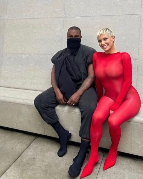 Bianca Censori kiedyś, czyli gdy wyszła za Kanye Westa. Oboje siedzą na łwce na tle betonowej ściany. Ona ma krótkie, blond włosy i czerwony kombinezon. On ma czarne spodnie, top i maskę na twarzy.