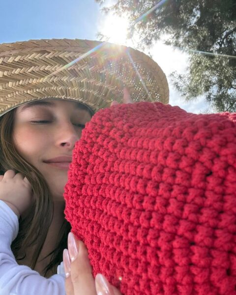 Zdjęcie przedstawiające celebrytkę Hailey Bieber ze słomianym kapeluszem na głowie. Trzyma w ramionach córeczkę ubraną w czerwoną czapeczkę. Widać fragment paznokci Hailey Bieber, które zrobiły furorę: glazed donut nails.