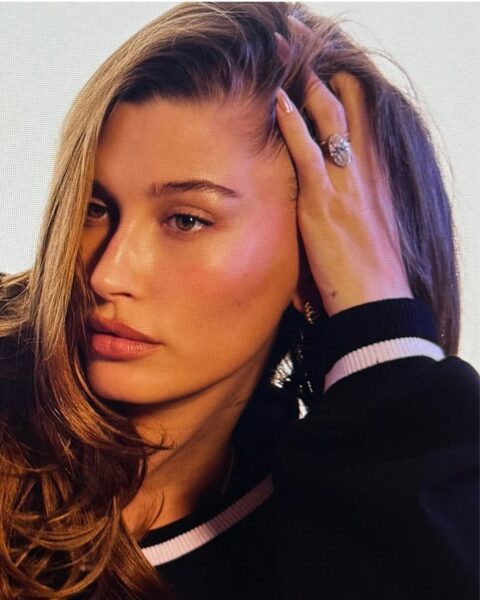 Zdjęcie przedstawiające manicure w stylu Hailey Bieber. Na zdjęciu jest celebrytka, która opiera się o dłoń, na której widać długie paznokcie glazed donut. Ubrana jest w czarną bluzę z białą lamówką przy dekolcie. Włosy ma zaczesane na jedną stronę.