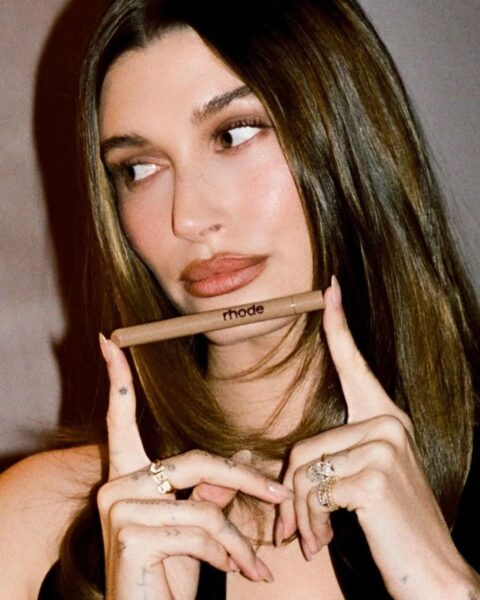 Zdjęcie przedstawiające celebrytkę Hailey Bieber. Ma ciemne, rozpuszczone włosy i ubrana jest w czarny top na ramiączkach i dużym dekoltem. W rękach trzyma konturówkę swojej marki Rhode. Na palcach ma mnóstwo złotych pierścionków.