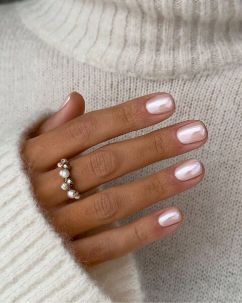 Zdjęcie przedstawiające dłoń i manicure w stylu Hailey Bieber, czyli glazed donut nails w kolorze różowym na krótkiej, kwadratowej płytce. Dłoń pozuje na tle kremowego swetra. Na palcu środkowym widać pierścionek z perełkami.