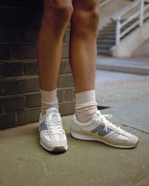 Zdjęcie skadrowane na nogi i buty New Balance w jasnym, beżowym kolorze z niebieski logo. Nosi je osoba w krótkich spodenkach, oparta o ceglaną ścianę.