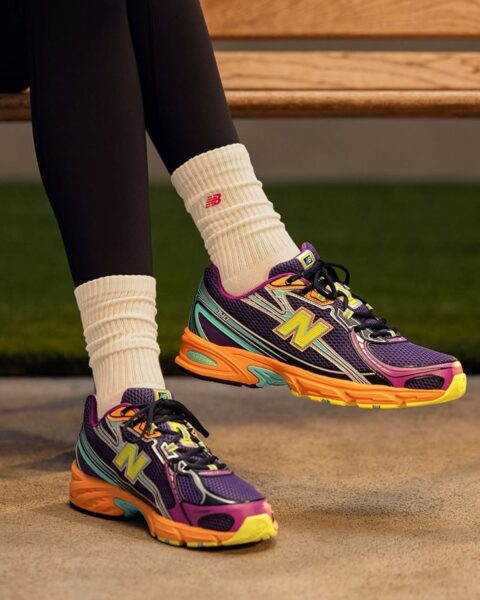 Zdjęcie skadrowane na nogi kobiety w czarnych legginsach, z kremowymi skarpetkami, siedzącą na drewnianej ławce, a która ma na nogach modne sneakersy New Balance na wiosnę 2025 w kolorze fioletowo-pomarańczowym.