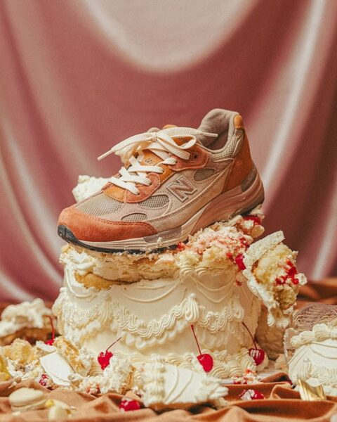 Artystyczne zdjęcie butów New Balance 992 w kolorze brzoskwiniowym ułożonych na torcie urodzinowym. W tle widać satynowe, kremowe kotary.