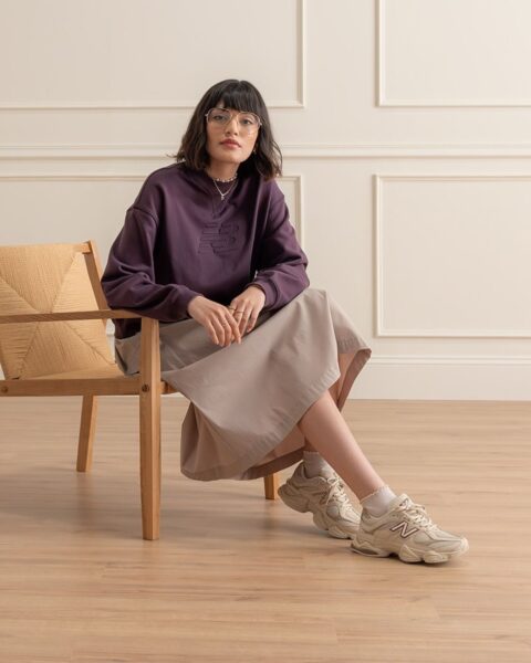Zdjęcie kobiety ubranej w śliwkową bluzę, beżową spódnicę oraz beżowe sneakersy New Balance. Ma na nosie okulary korekcyjne, a na szyi naszyjniki. Włosy krótkie, ciemne, z grzywką. Siedzi na drewnianym krześle w przestronnym pomieszczeniu z ozdobną sztukaterią na kremowych ścianach.