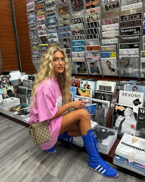 Kobieta z długimi falowanymi blond włosami pozuje przy stojaku z czasopismami. Ma na sobie różową, oversize'ową koszulkę oraz niebieskie bokserskie sneakersy adidas.