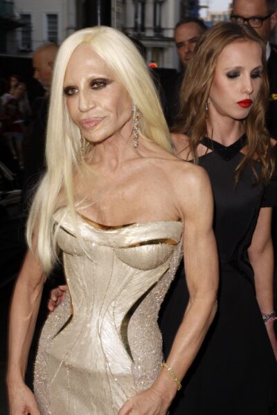 Donatella Versace. Kobieta w blond włosach pozuje w złotej sukni z gorsetową górą i ciemnym makijażu. W tle widać innych uczestników wydarzenia.