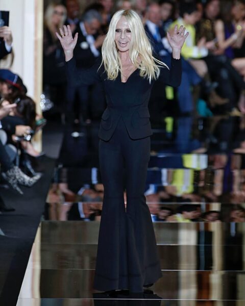 Donatella Versace w czarnym total looku. Macha do widowni po zakończonym pokazie mody.