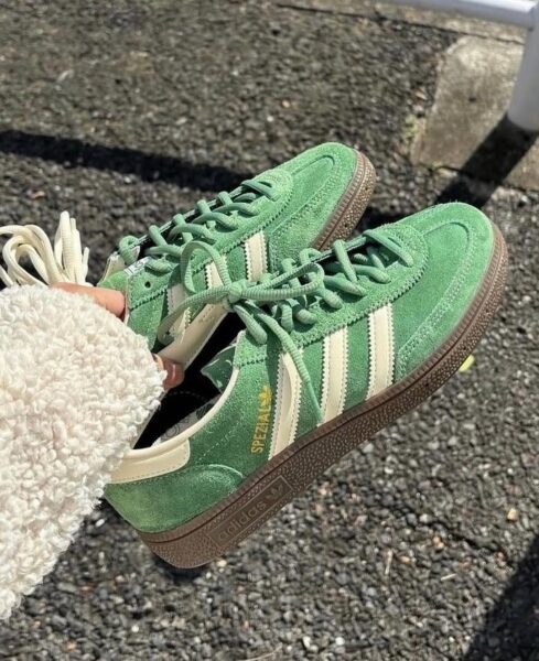 Na zdjęciu widoczna jest para zielonych zamszowych sneakersów Adidas Spezial, które są modne w kwietniu 2025. Buty mają charakterystyczne trzy białe paski po bokach i brązową gumową podeszwę. Sneakersy są trzymane przez kobietę mającą na sobie jasny, puszysty sweter.