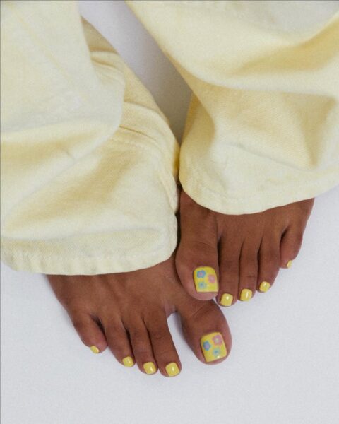 Radosny pedicure w żółtym kolorze z namalowanymi kolorowymi kwiatkami na dużych palcach, zestawiony z żółtymi spodniami – idealny wybór na słoneczne dni.