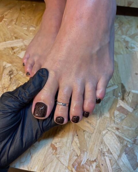 Brokatowy pedicure w kolorze gorzkiej czekolady, prezentowany na tle drewnianej podłogi – stylizacja z eleganckim pierścionkiem i profesjonalnym wykończeniem.