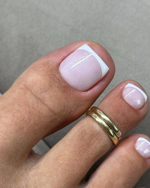 Zbliżenie na paznokieć z delikatnym french pedicure w wersji glamour – mlecznoróżowa baza z cienką, białą końcówką i subtelnym połyskiem, idealna stylizacja na lato.