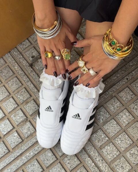 Zdjęcie przedstawia dłonie kobiety z ciemnymi paznokciami, licznymi pierścionkami i bransoletkami. Kobieta nosi białe sneakersy Adidas Taekwondo z czarnymi paskami. Sneakersy są połączone z ozdobnymi skarpetkami z falbankami.