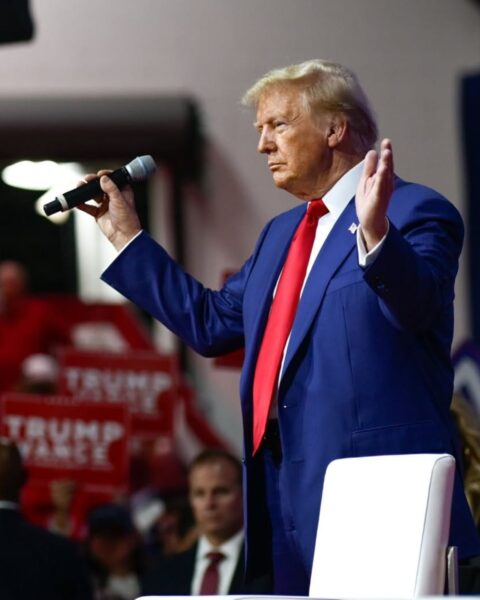 Zdjęcie Donalda Trumpa podczas swojego wiecu wyborczego. Ubrany jest granatowy garnitur, białą koszulę i czerwony krawat. Rozkłada ręce, a w jednej trzyma mikrofon.