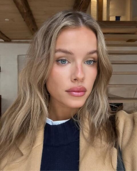 Zdjęcie młodej kobiety, która zrobiła sobie selfie. Ma blond włosy, rozpuszczone na ramiona. Ubrana jest w granatowy sweter i beżowy płaszcz. Ma na ramieniu torebkę Pozuje w efektownym mieszkaniu, widać za nią schody i sufit obity deskami.