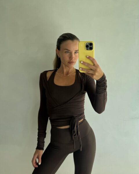 Zdjęcie kobiety, która zrobiła sobie selfie. Ubrana jest w strój do pilatesu w kolorze brązowym składający się z topu, bluzki oraz legginsów. Ma związane włosy.