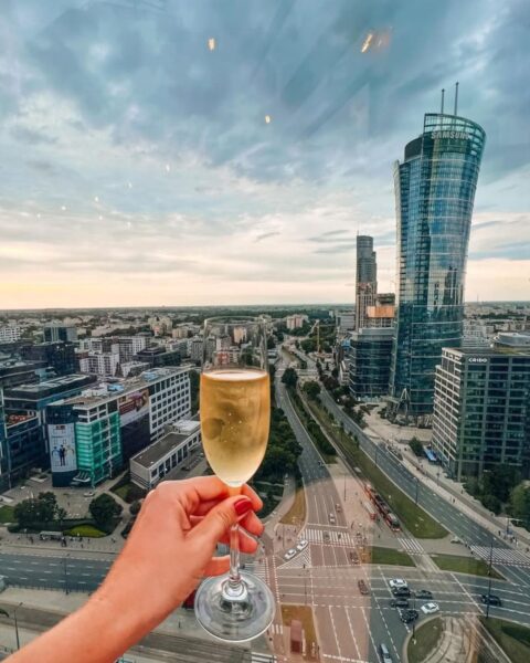 Najlepsze bary z rooftopami Warszawa: zdjęcie z widokiem na centrum Warszawy z biurowcami. Widać na pierwszym planie dłoń wyciągniętą z kieliszkiem.