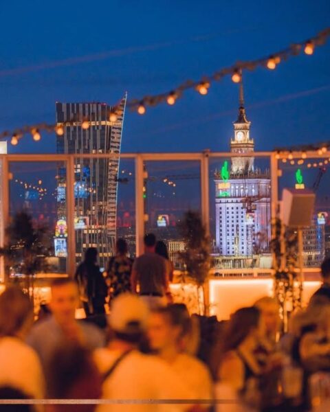 Zdjęcie przedstawiające klub nocny na dachu budynku w Warszawie. To rooftop z widokiem na panoramę miasta. Na zdjęciu widać bawiących się ludzi, szklaną balustradę i rozciągnięte nad ich głowami girlandy.