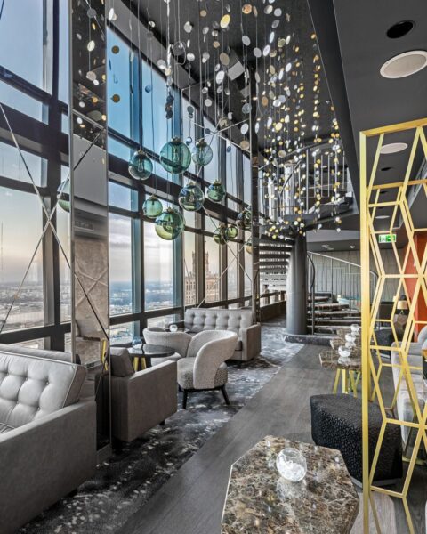 Zdjęcie przedstawiające bar na rooftopie w nowoczesnym stylu. Widać wnętrze z wysokim sufitem, dużymi przeszkleniami, kanapami, fotelami, marmurowymi stolikami oraz wiszącymi, szklanymi lampami. Wszystko utrzymane jest w chłodnym, szaro-srebrnym stylu i przełamane kolorem zieleni lub żółci.
