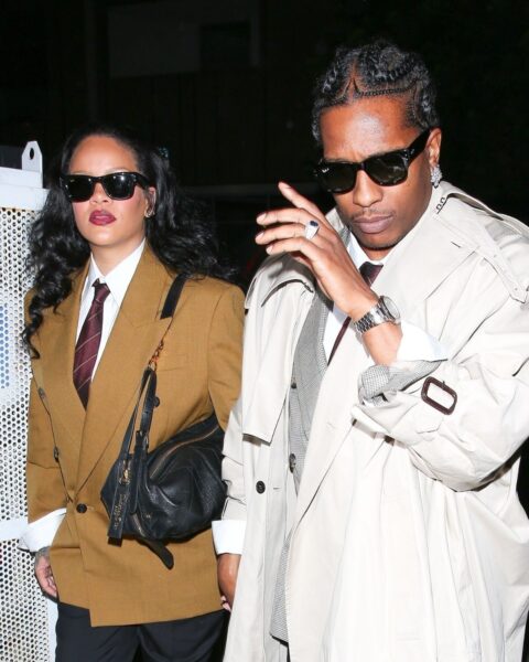 Rihanna i ASAP Rocky. Rihanna ma na sobie białą koszulę, krawat, karmelową marynarkę oraz czarne spodnie, torebkę czarną oraz okulary przeciwsłoneczne na nosie. ASAP Rocky ma na sobie garnitur, a do tego szeroki, kremowy trencz i okulary przeciwsłoneczne. Idą wieczorem ulicą.