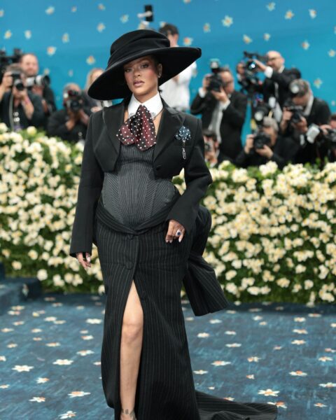 Rihanna w trzeciej ciąży podczas gali Met Gala 2025. Ma na sobie czarną kreację przypominającą garnitur z szarym gorsetem. Ma do tego kapelusz na głowie, krawat, biały kołnierzyk. Stoi przed schodami przed wejściem do Metropolitan Museum of Art.