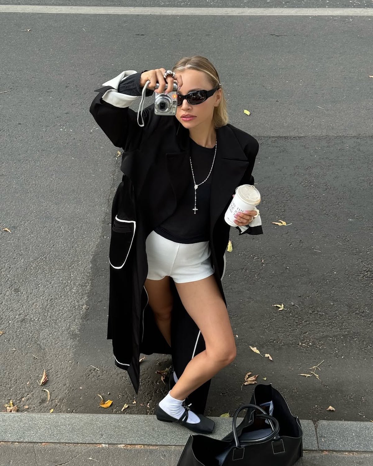 Stylowo ubrana kobieta stoi na chodniku, trzymając w jednej ręce aparat fotograficzny skierowany w stronę obiektywu, a w drugiej kubek z kawą na wynos. Ma na sobie długi czarny płaszcz z białymi detalami, czarną koszulkę, białe krótkie spodenki, białe skarpetki i czarne buty typu Mary Jane. Uzupełnieniem stylizacji są okulary przeciwsłoneczne, naszyjnik z krzyżykiem i duża czarna torba stojąca obok niej na ziemi.