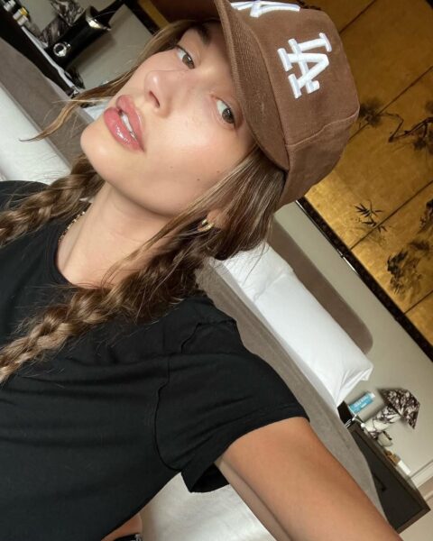 Hailey Bieber w brązowej czapce z daszkiem, dwóch warkoczach i czarnej koszulce. Celebrytka ma delikatny makijaż, pozuje na tle łóżka.