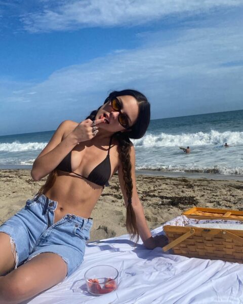 Olivia Rodrigo siedzi na plaży w bikini i jeansowych szortach. Piosenkarka pozuje na tle morza w dwóch długich warkoczykach i okularach przeciwsłonecznych.