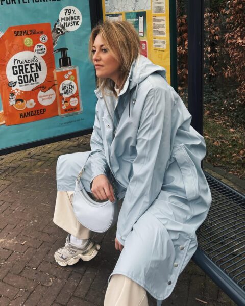 Blondynka siedzi na przystanku. Ma na sobie długi jasnoniebieski płaszcz przeciwdeszczowy z kapturem, beżowe spodnie i sneakersy New Balance.