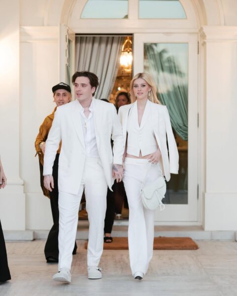 Brooklyn Beckham i Nicola Peltz w eleganckich białych garniturach na tle rezydencji – Para w dopasowanych stylizacjach typu total white wychodzi z pałacowego budynku, trzymając się za ręce, emanując hollywoodzkim szykiem i nowoczesną interpretacją klasyki.