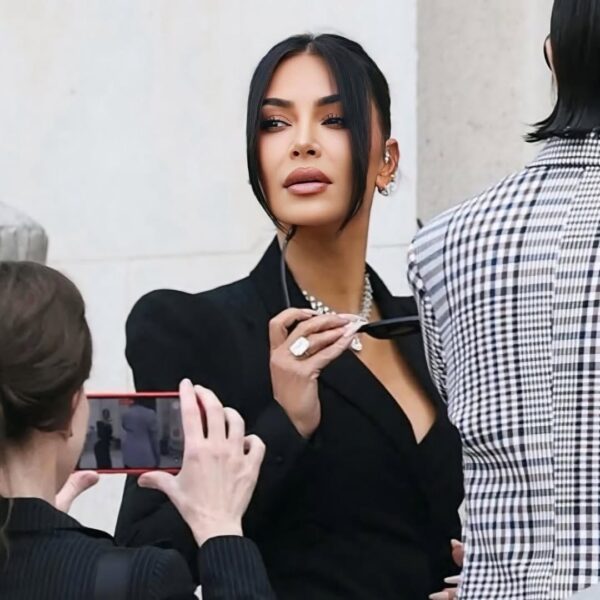 Kim Kardashian proces: Kobieta spoglądająca przez ramię, ubrana w czarną, taliowaną suknię z baskinką, z włosami spiętymi w kok, trzymająca rękę w powietrzu – elegancka poza w stylu red carpet.