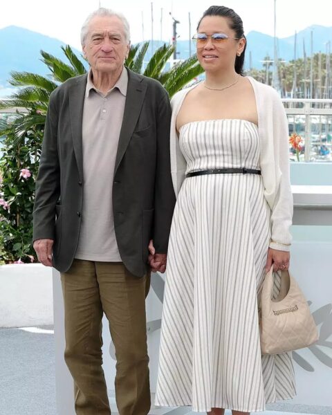 Robert De Niro i Tiffany Chen pozują razem podczas festiwalu filmowego na tle palm i przystani. Aktor ubrany w lnianą koszulę i marynarkę, Tiffany w biało-szarą sukienkę w paski, narzucony kardigan i trzymająca jasną torebkę. Oboje uśmiechnięci i eleganccy w luźnej, letniej stylizacji.