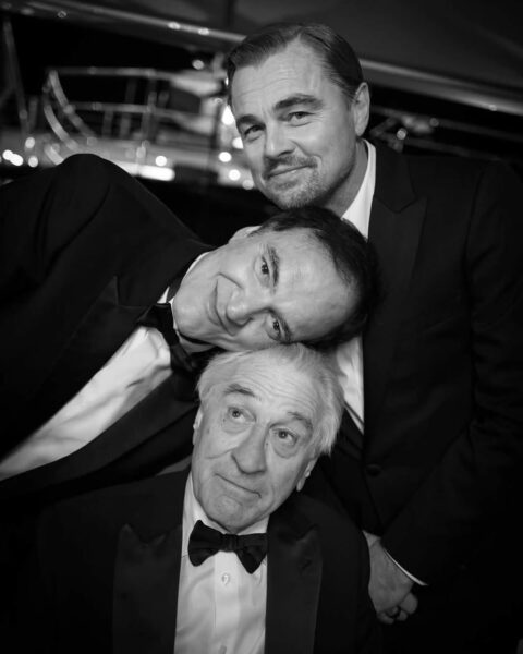Czarno-białe zdjęcie przedstawiające Roberta De Niro, Quentina Tarantino i Leonardo DiCaprio w ciepłym, przyjacielskim uścisku. Tarantino opiera głowę na ramieniu De Niro, a DiCaprio obejmuje ich z tyłu. Fotografia emanuje nostalgiczną atmosferą i celebracją wieloletnich przyjaźni w świecie kina.