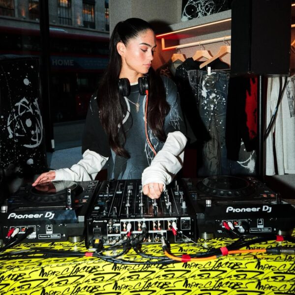 DJ-ka grająca set na imprezie w sklepie odzieżowym – Młoda kobieta z długimi ciemnymi włosami i słuchawkami na szyi operuje konsoletą DJ-ską w otoczeniu ubrań i miejskiego neonu za oknem, prezentując energię muzycznego wydarzenia.