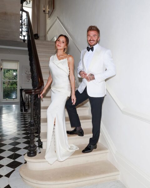 David i Victoria Beckham pozują na schodach w eleganckich wieczorowych strojach – Ona w efektownej białej sukni z rozcięciem i upięciem na jedno ramię, on w białym smokingu z czarną muchą – oboje promienieją klasą i stylową nonszalancją.