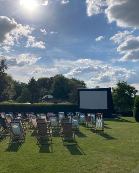 Zdjęcie przedstawiające kino plenerowe na łące otoczonej żywopłotem i drzewami. Widać wielki ekran a przed nim rząd krzeseł.