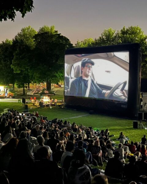 Zdjęcie przedstawiające kino plenerowe. Widać wielki ekran z filmem, na łące, naprzeciwko rzędy widzów. W tle widać park z food truckami, parasolami i miejscem do picia. 