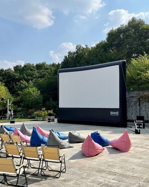 Zdjęcie przedstawiające kino plenerowe na placu z płytkami. Widać wielki ekran, a przed nim rzędy siedzeń i krzeseł. W tle widać drzewa. 
