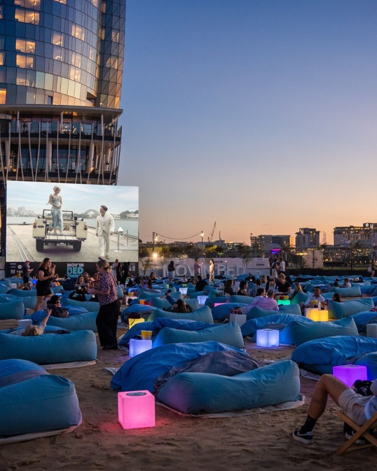 Zdjęcie ilustrujące kina letnie w Warszawie, czyli kino wystawiane na zewnątrz. Na zdjęciu jest plaża pełna miękkich leżaków w kolorze niebieskim, wielki ekran z filmem oraz panorama miasta i wysoki, szklany budynek.
