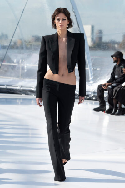 Modelka na wybiegu w eleganckim czarnym garniturze z ekstremalnie niskim stanem i odsłoniętym torsem, z pokazu Alexander McQueen Pre-Spring 2023. Minimalistyczna, ale szokująca stylizacja redefiniująca kobiecą sylwetkę w kontekście mody wieczorowej.