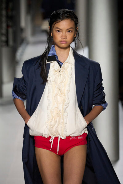 Modelka na wybiegu Miu Miu wiosna-lato 2024 w granatowym oversize’owym płaszczu, kremowej bluzie z falbanami i czerwonych sportowych majtkach z logo Miu Miu. Stylizacja łączy estetykę akademicką z odważnym, bieliźnianym twistem.