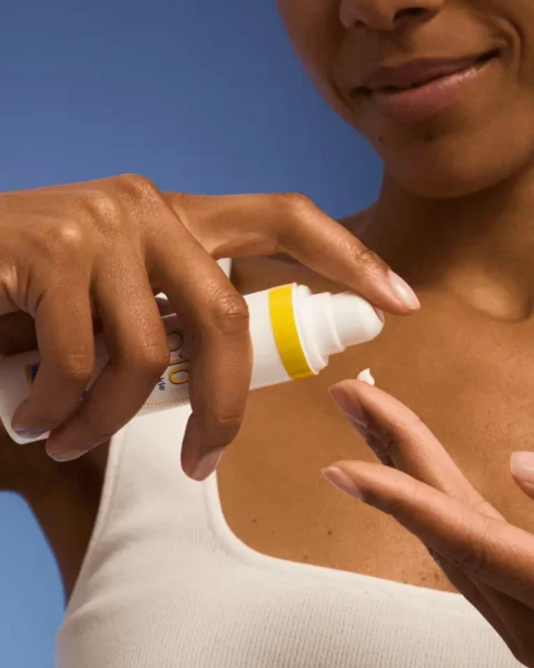 Zbliżenie na dłoń kobiety aplikującej dzienny fluid z filtrem SPF 50 marki Nivea z serii Q10. Produkt w białej tubce z żółtym akcentem, idealny do codziennej ochrony przeciwsłonecznej i pielęgnacji skóry z oznakami starzenia. Tło stanowi błękitne studio, a kobieta nosi biały top.