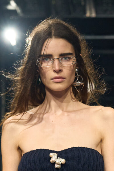 Zbliżenie na modelkę z pokazu Prada FW25 w czarnej, dzianinowej sukni bez ramiączek z aplikacją z pereł. Na twarzy wyraziste okulary z ozdobną metalową ramką, dodające futurystycznego klimatu całości.