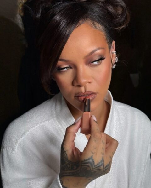Rihanna straciła fortunę. Na zdjęciu jest Rihanna ubrana w biały szlafrok. Włosy ciemne, spięte w kok. Maluje sobie usta szminką swojej marki Fenty Beauty.