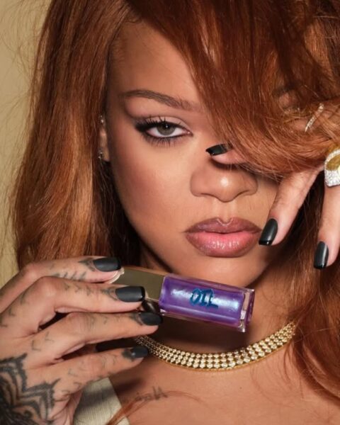 Rihanna w rudych włosach z długą grzywką na czole. Ubrana jest w beżowy top, ma złotą kolię z cyrkoniami, pomalowane na czarno paznokcie, masywny pierścień złoty na palcu. W ręce trzyma olejek do ust Fenty Beauty, swojej marki.