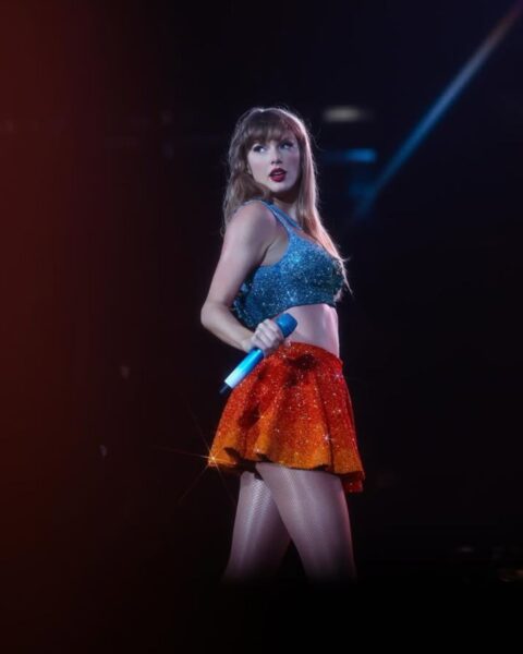 Taylor Swift w trakcie swojego koncertu. Ma na sobie niebieski top oraz pomarańczową minispódniczkę wyszywane cekinami. W ręce trzyma mikrofon. Stoi półbokiem, obraca się do publiczności. 