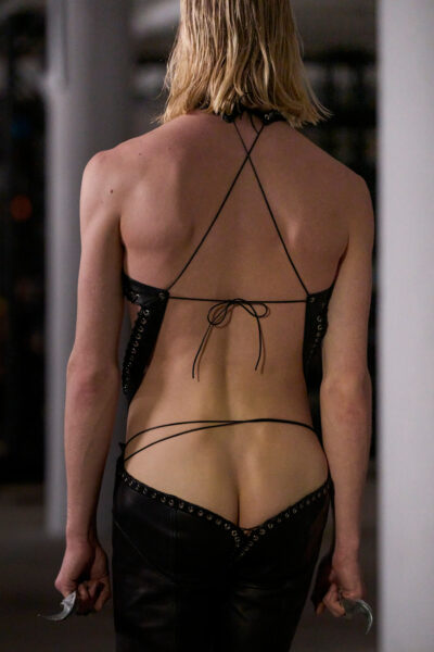 Model w czarnych, skórzanych spodniach z bardzo niskim stanem i topie typu string-back z wiązaniami, odsłaniający plecy i pośladki. Stylizacja z pokazu Ludovica de Saint Sernina ukazuje modę jako przestrzeń dla ekspresji płci i zmysłowości.