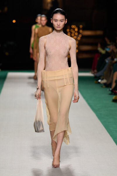 Modelka na wybiegu w przezroczystym body i spódnicy o nietypowej, wysokiej talii z kolekcji Victoria Beckham Spring 2025. Stylizacja eksploruje temat cielesności i geometrii poprzez delikatne, półprzezroczyste tkaniny.